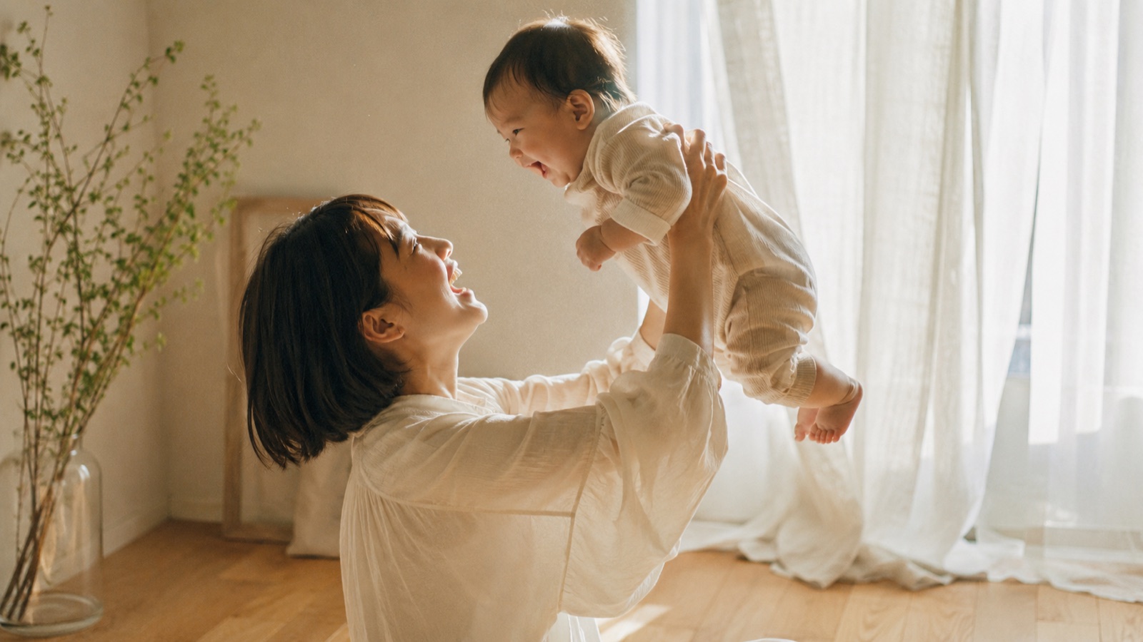 窓辺のやわらかな光の中で、赤ちゃんを抱き上げて笑い合う母親